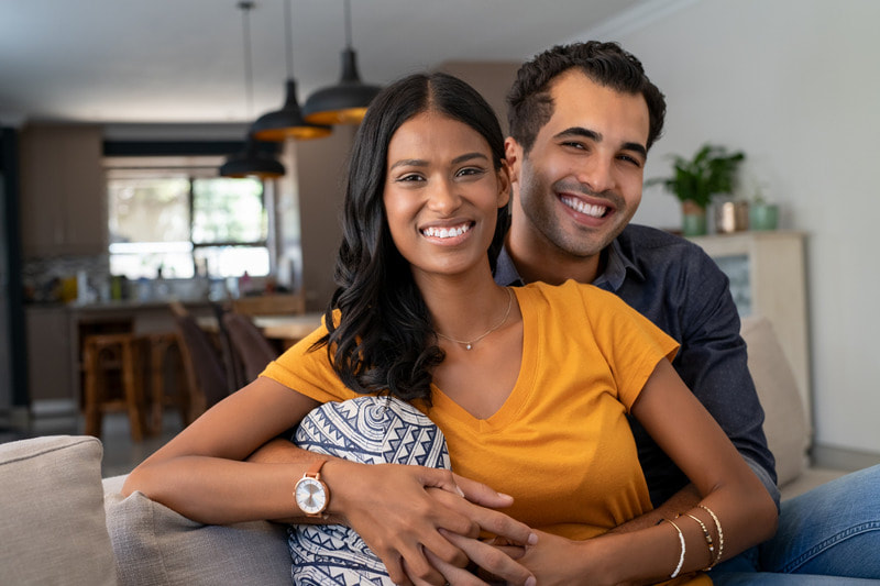 Multicultural couple cuddling on a couch in their new home at Pinnacle Hervey Bay. The Bengali Indian wife is late 20's and wearing a mustard yellow shirt.  The husband is Sri Lankan and proud that they have purchased their first property.